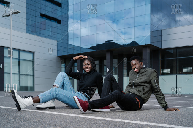 Full body young black man and woman in casual clothes smiling and looking at camera while sitting on asphalt ground on parking lot outside contemporary building on city street