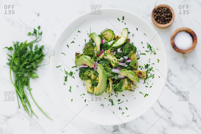 From above of round white plate with served pile of avocado salad with zucchini and broccoli garnished with chopped red onion and parsley