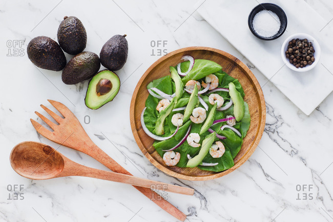 Flat lay of round wooden plate with salad of green and avocado garnished with red onion and shrimps composed with ingredients on white marble