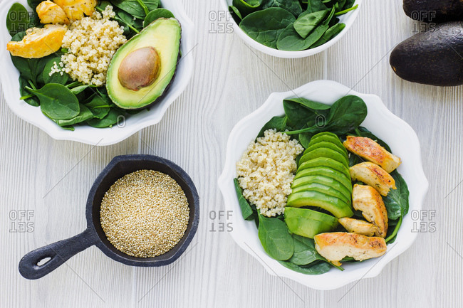 Top view of white bowls with green spinach garnished with couscous and avocado with friend chicken on wooden table