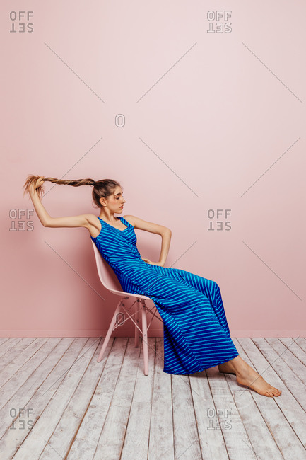 Side view of young woman in blue dress barefoot looking at camera holding ponytail while sitting in chair and looking away on minimalist pink background