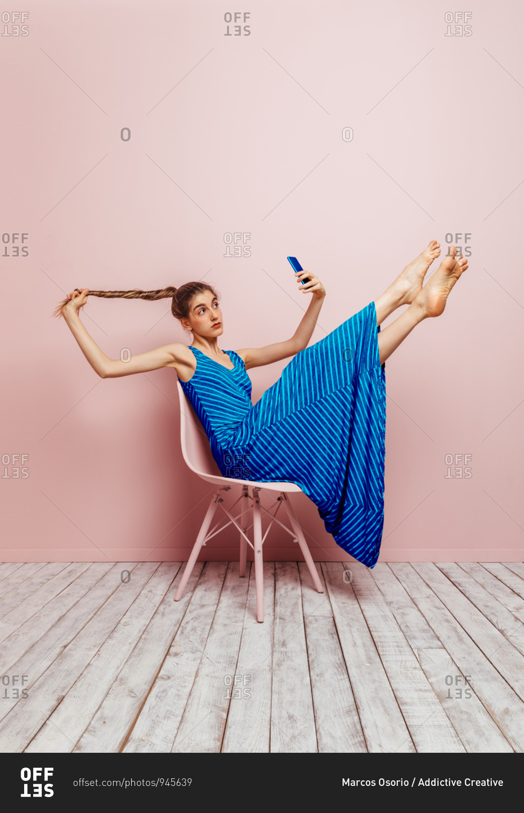 Side view of woman sitting in a chair lifting legs up barefoot looking at mobile phone while taking a selfie holding ponytail up on pink background