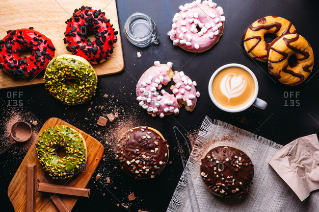Flat lay of various doughnuts with sweet toppings and chocolate bars composed with cup of cappuccino on black table