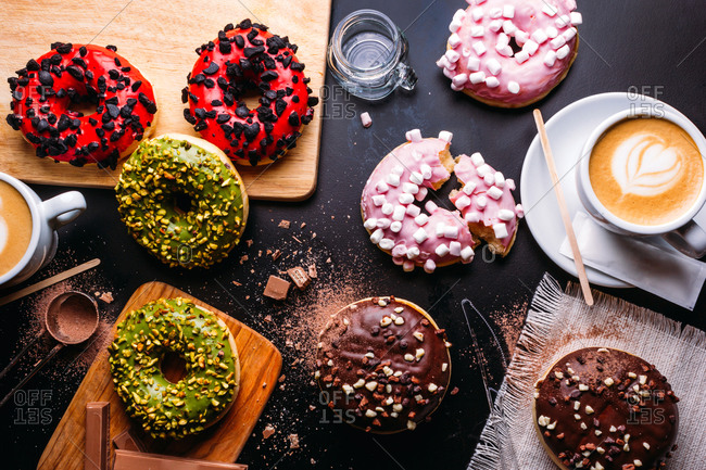 Flat lay of various doughnuts with sweet toppings and chocolate bars composed with cup of cappuccino on black table