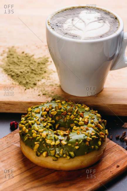 High angle of delicious doughnut with green topping and chopped nuts placed on wooden board with cup of aromatic cappuccino