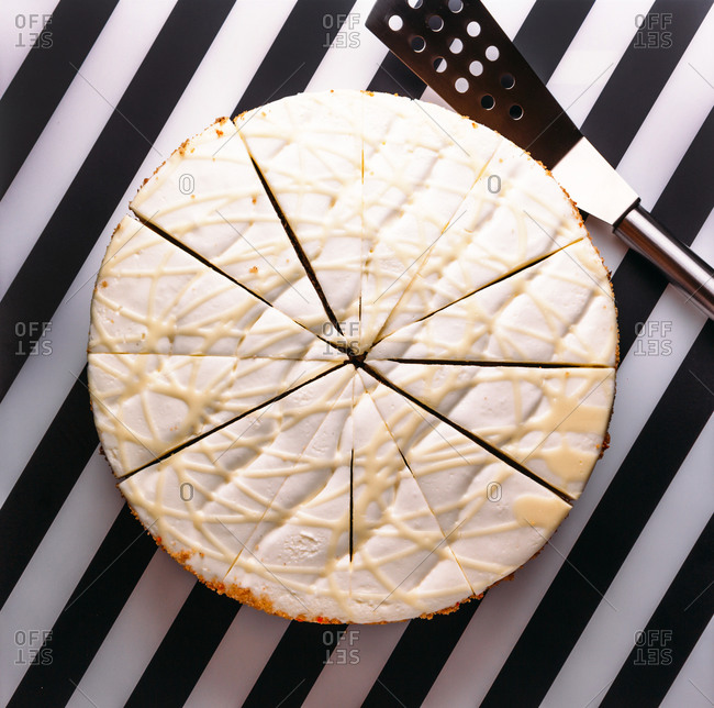 Top view of round cake with white cream and glaze cut in pieces for serving placed on black and white striped surface