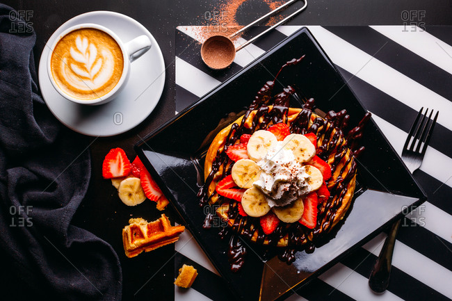 Top view of cappuccino in white mug on table with plate of round waffle with banana and strawberry topped with chocolate sauce and whipped cream