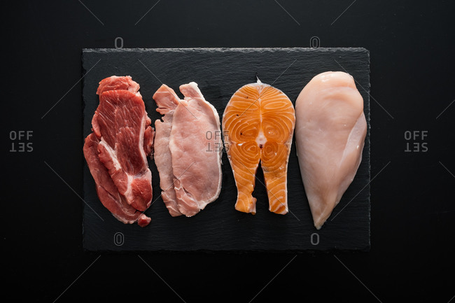 From above fresh raw fillet of beef and pork with chicken and fish pieces on wooden cutting board as composition of protein rich food against black background