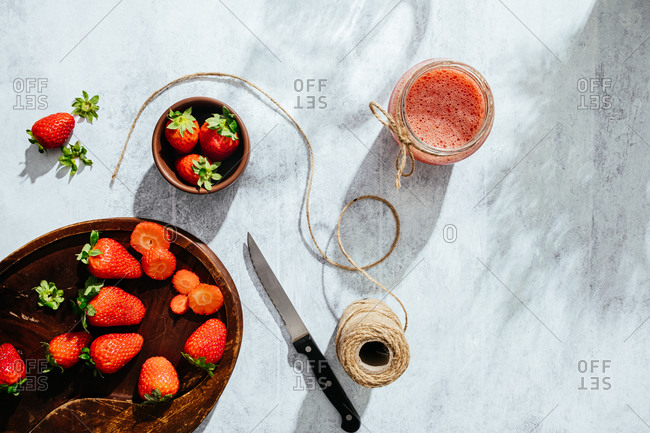 From above composition with fresh homemade strawberry juice in glass jar wrapped with twine placed on marble surface with whole berries and knife