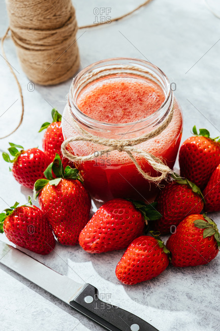 Composition with fresh homemade strawberry juice in glass jar wrapped with twine placed on marble surface with whole berries and knife