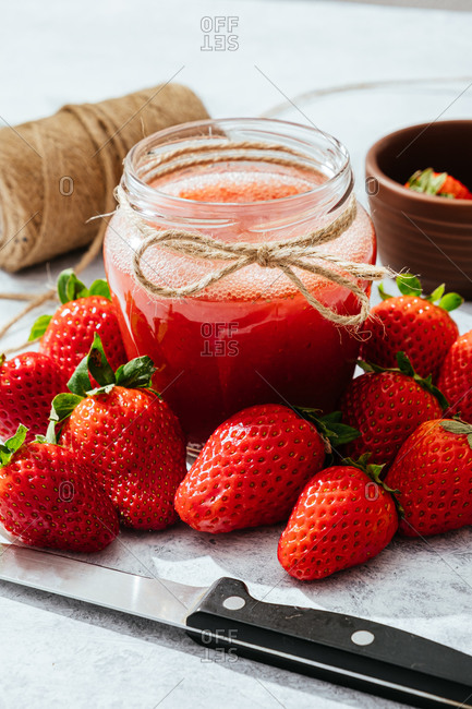 Composition with fresh homemade strawberry juice in glass jar wrapped with twine placed on marble surface with whole berries and knife