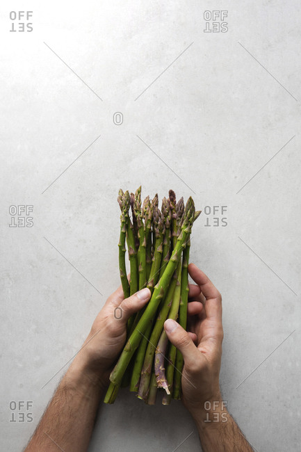 Top view of crop anonymous chef holding bunch of fresh green asparagus while preparing food at gray marble table in kitchen