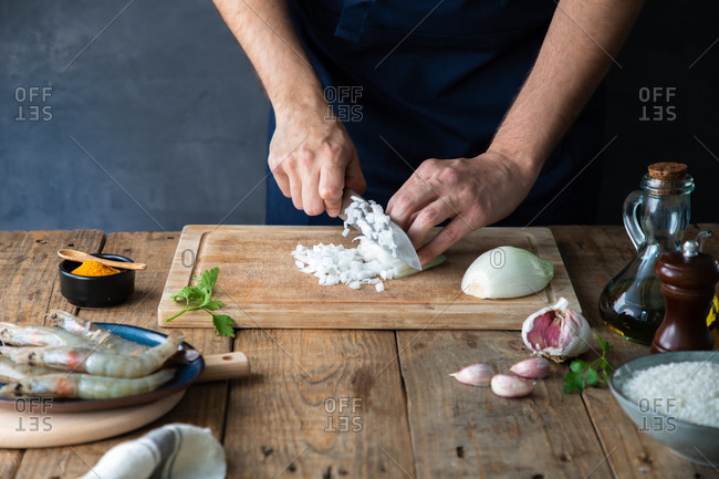 From above faceless chef cutting onion on cutting board while preparing appetizing dish at wooden table with bowl of rice and raw shrimps in composition with other ingredients at home