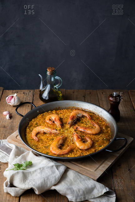 High angle of big metal pan of while appetizing paella with roasted shrimps on rustic table in composition with vintage bottle of oil and towel in kitchen at home