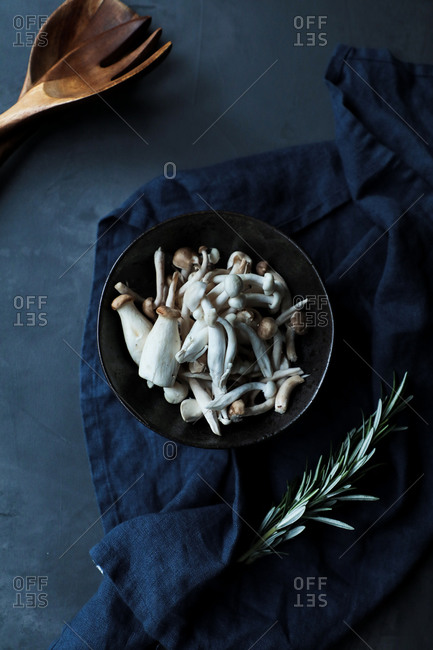 Top view of bowl with uncooked mushrooms and fresh green rosemary stems on  dark blue towel in arrangement with wooden spoon and fork on black table in rustic kitchen