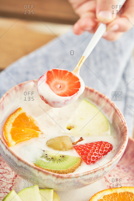 From above of crop unrecognizable female hands having healthy breakfast and eating delicious yogurt with sliced fresh assorted fruits served on wooden table