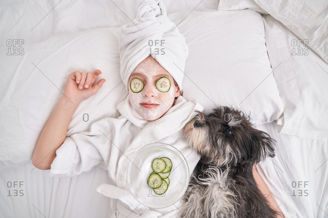 Top view of child in white bathrobe and towel turban with facial mask and cucumber slices on eyes lying on bed with fluffy dog while relaxing during spa procedure at home