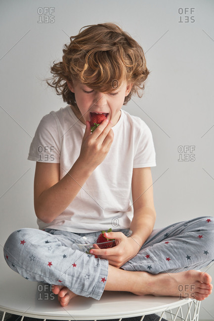 Boy in sleepwear biting sweet strawberry while sitting cross legged on table against gray wall at home