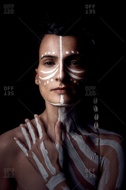 Portrait of naked tender beautiful brunette Native American woman with white striped painted on body covering breast standing in dark on black background looking at camera