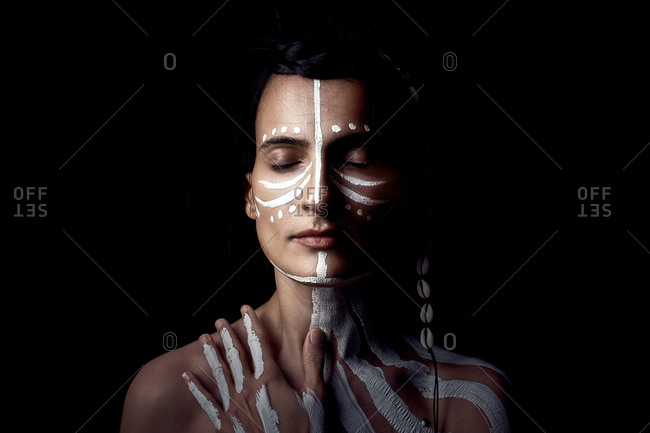 Portrait of naked tender beautiful brunette Native American woman with white striped painted on body covering breast standing in dark on black background with closed eyes