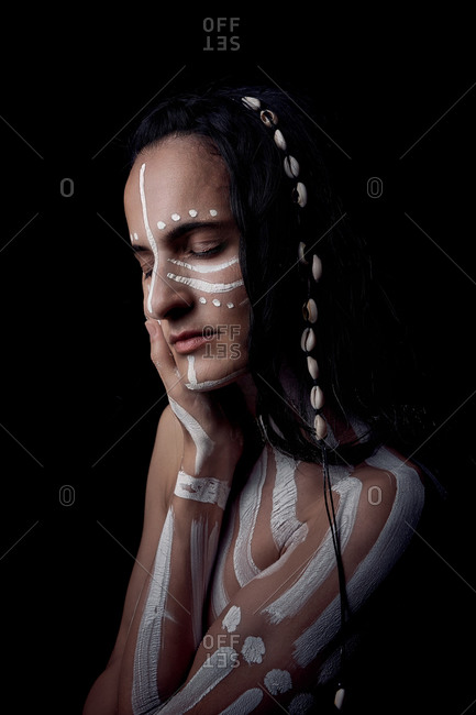 Side view portrait of naked tender beautiful brunette Native American woman with white striped painted on body covering breast standing in dark on black background with closed eyes