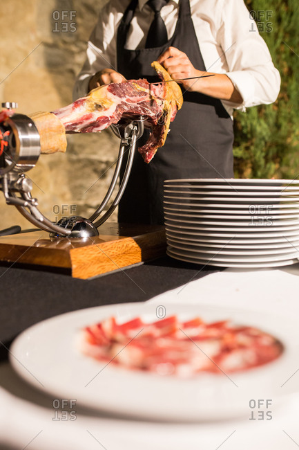 Crop unrecognizable person hand on apron cutting a whole dry-cured ham leg on a black background