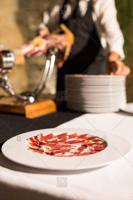 Crop unrecognizable person hand on apron cutting a whole dry-cured ham leg on a black background