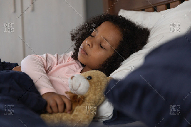 African American girl sleeping in her father bedroom and embracing a teddy bear, during social distancing at home during quarantine lockdown.