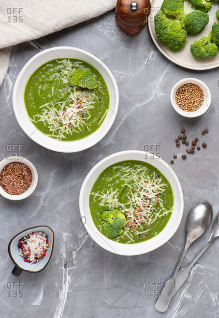 Overhead view of two bowls with green broccoli cream soup served with ground cheese parmesan and linen seeds