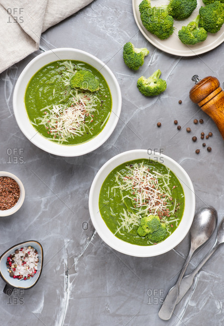 Overhead view of two bowls with green broccoli cream soup served with ground cheese parmesan and linen seeds