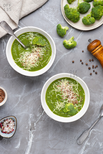 Overhead view of two bowls with green broccoli cream soup served with ground cheese parmesan and linen seeds