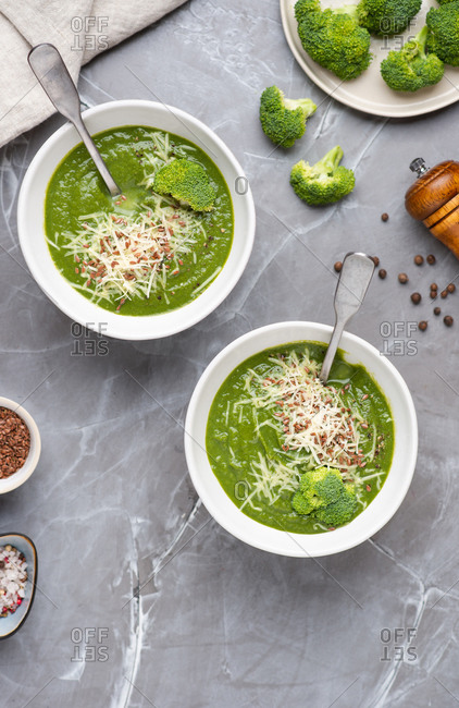 Overhead view of two bowls with green broccoli cream soup served with ground cheese parmesan and linen seeds