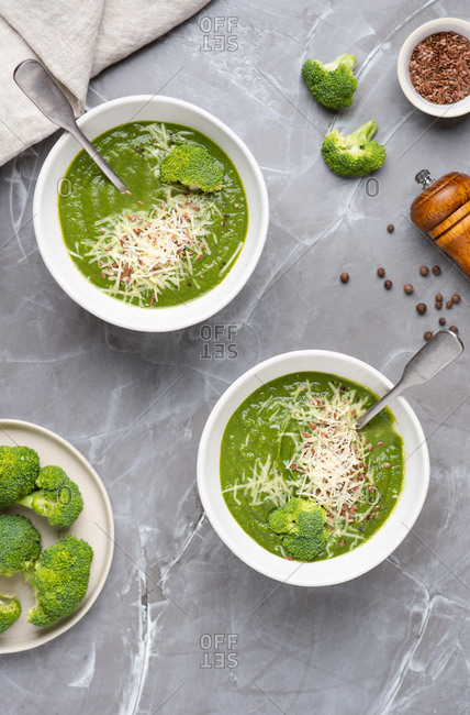 Overhead view of two bowls with green broccoli cream soup served with ground cheese parmesan and linen seeds
