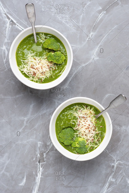 Overhead view of two bowls with green broccoli cream soup served with ground cheese parmesan and linen seeds
