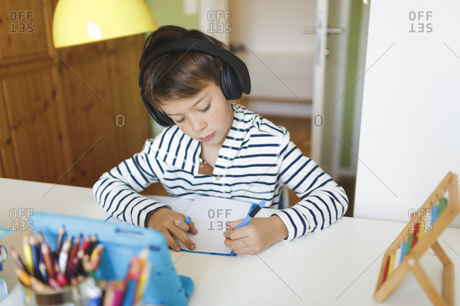 Boy doing homeschooling and writing on notebook- using tablet and headphones at home
