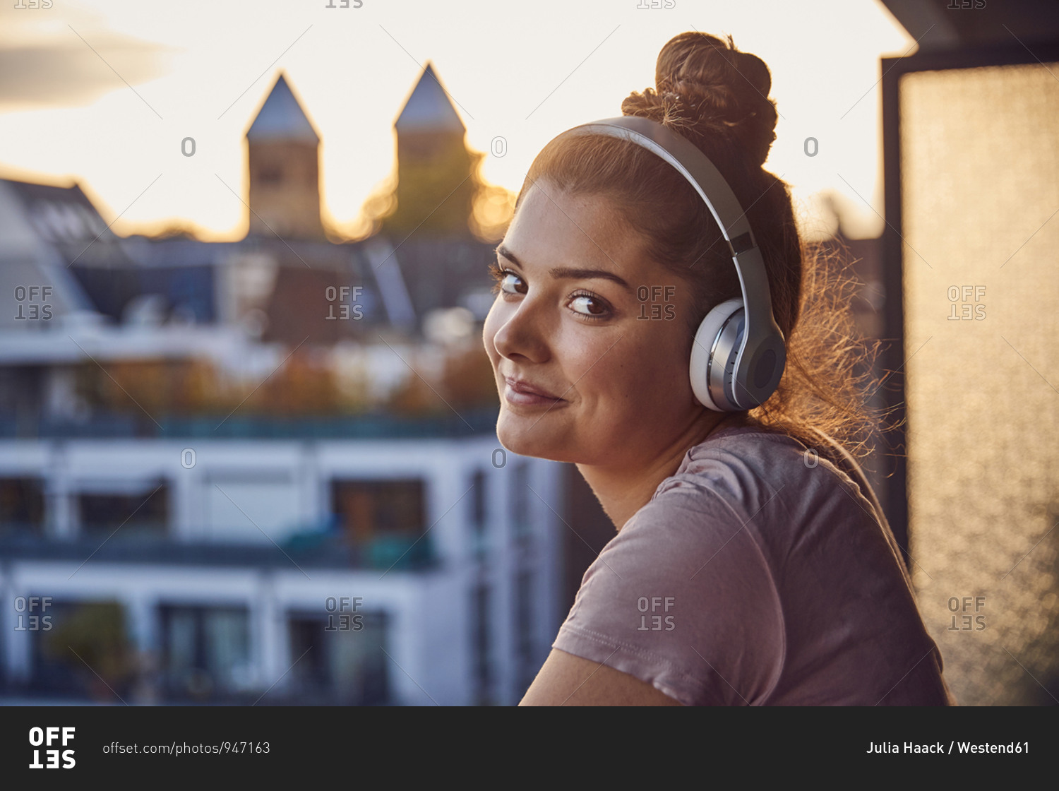 Portrait of smiling young woman with bun listening music with headphones on balcony in the evening
