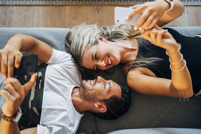 Happy couple lying on the couch taking selfies with smartphones while looking at each other