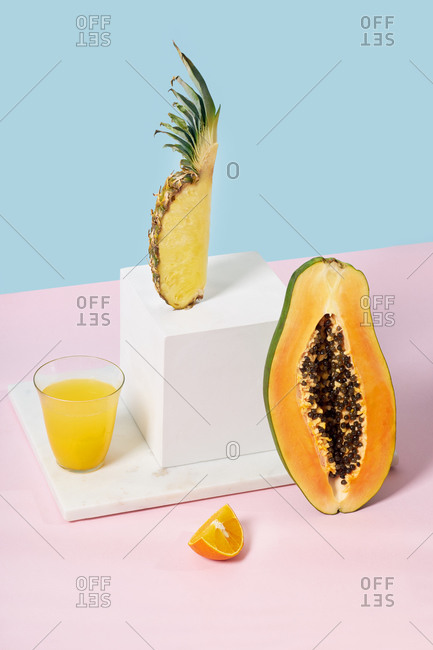 Still life with pineapple slice, white cube, papaya half and a glass of orange juice on colorful background