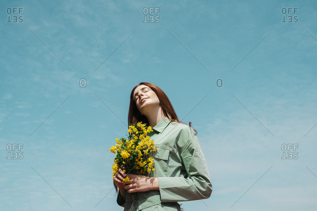 Portrait of redheaded young woman with eyes closed standing against sky holding bunch of yellows flowers