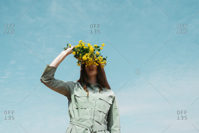 Redheaded young woman against sky covering eyes with bunch of yellow flowers