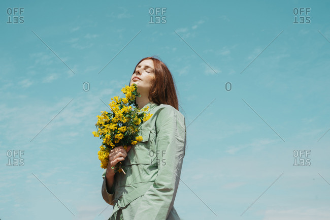 Portrait of redheaded young woman with eyes closed standing against sky holding bunch of yellow flowers