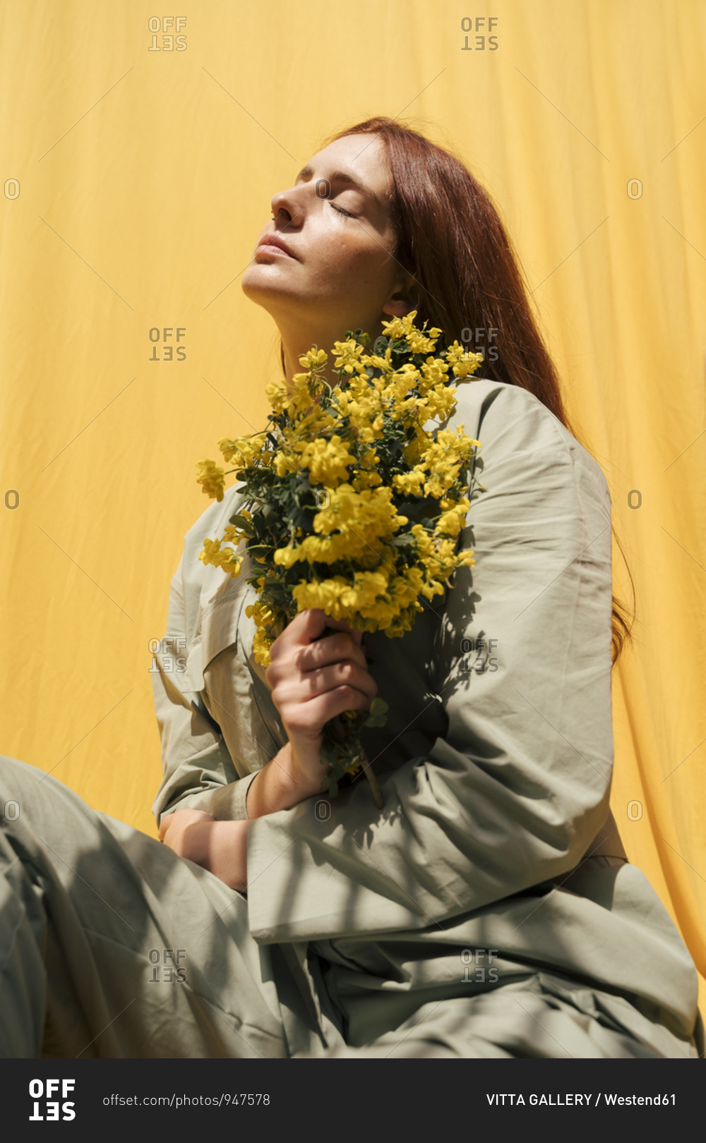 Portrait of redheaded woman with eyes closed holding bunch of yellow flowers against yellow background