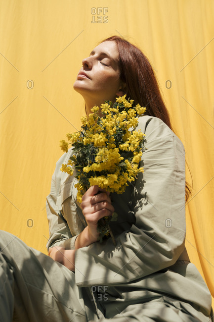 Portrait of redheaded woman with eyes closed holding bunch of yellow flowers against yellow background