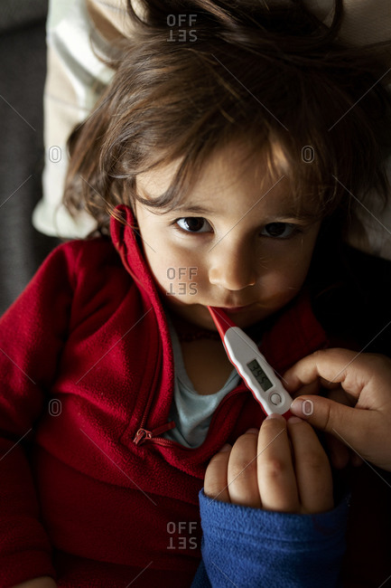 Portrait of sick girl lying on couch with digital thermometer- mother's hands