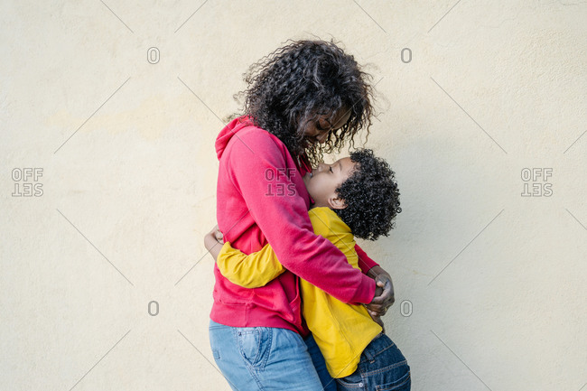 Mother and son dressed in colorful casual clothes hugging each other tenderly outside with a yellow wall in the background