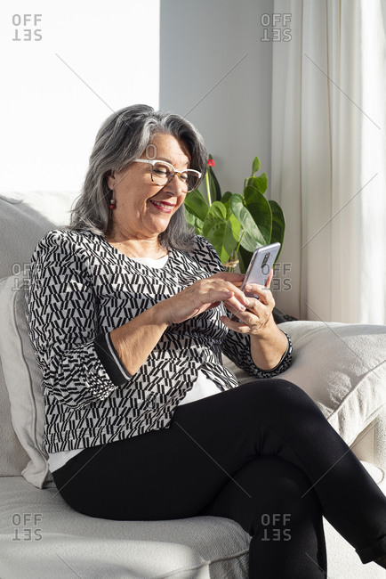 Smiling old lady sitting on a sofa and using a smartphone mobile
