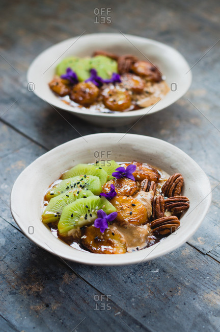 Warm oatmeal porridge with caramelized bananas, pecan nuts, tahini and kiwi on rustic wood surface