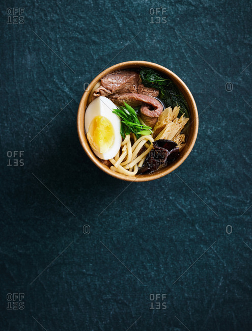 Minimalistic composition with Japanese beef ramen noodle soup in take away packaging on dark textile background
