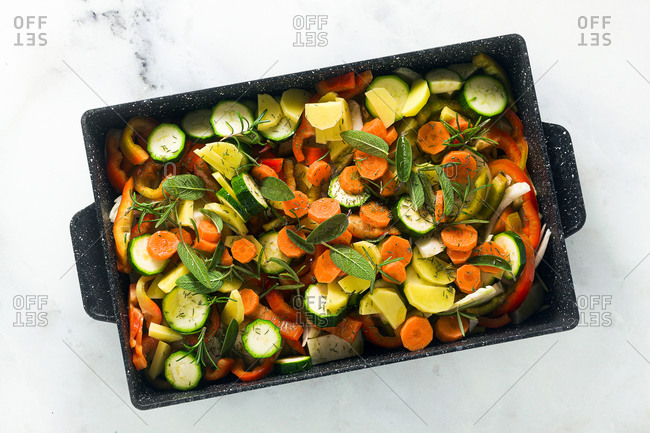 Various vegetables and aromatic herbs in a baking sheet, ready for baking on a white marble table, copy space. Top view.