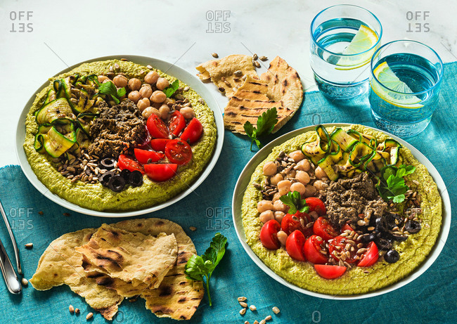 Home cooked hummus with fresh peas and stuffed with olive pate, grilled zucchini, olives and cherry tomato with toasted sunflower seeds and flat bread on the table covered blue textile, copy space.